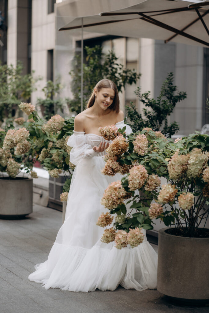 Side view of beautiful young man wearing wedding dress, posing outdoors. Attractive bride standing, smiling, looking at flowers, smiling. Concept of beauty and happiness.